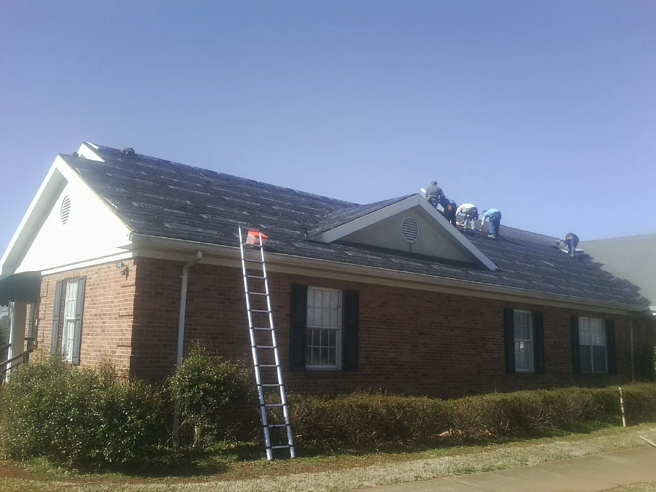 Crew tearing shingles off a brick ranch roof with a ladder set — Metal Roof Repair work by our crew in Mattituck