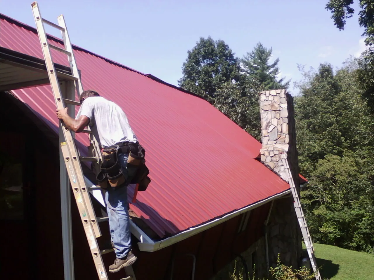 Roofer on a ladder beside a red metal roof and stone chimney — Soffit & Fascia Repair by Roofing Brothers in Mattituck